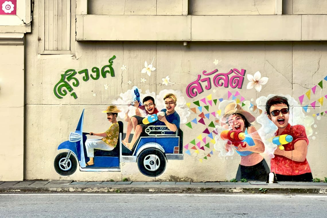 Peinture murale colorée de personnes souriantes dans un tuk-tuk jouant avec des pistolets à eau, entourées de fleurs et de fanions.