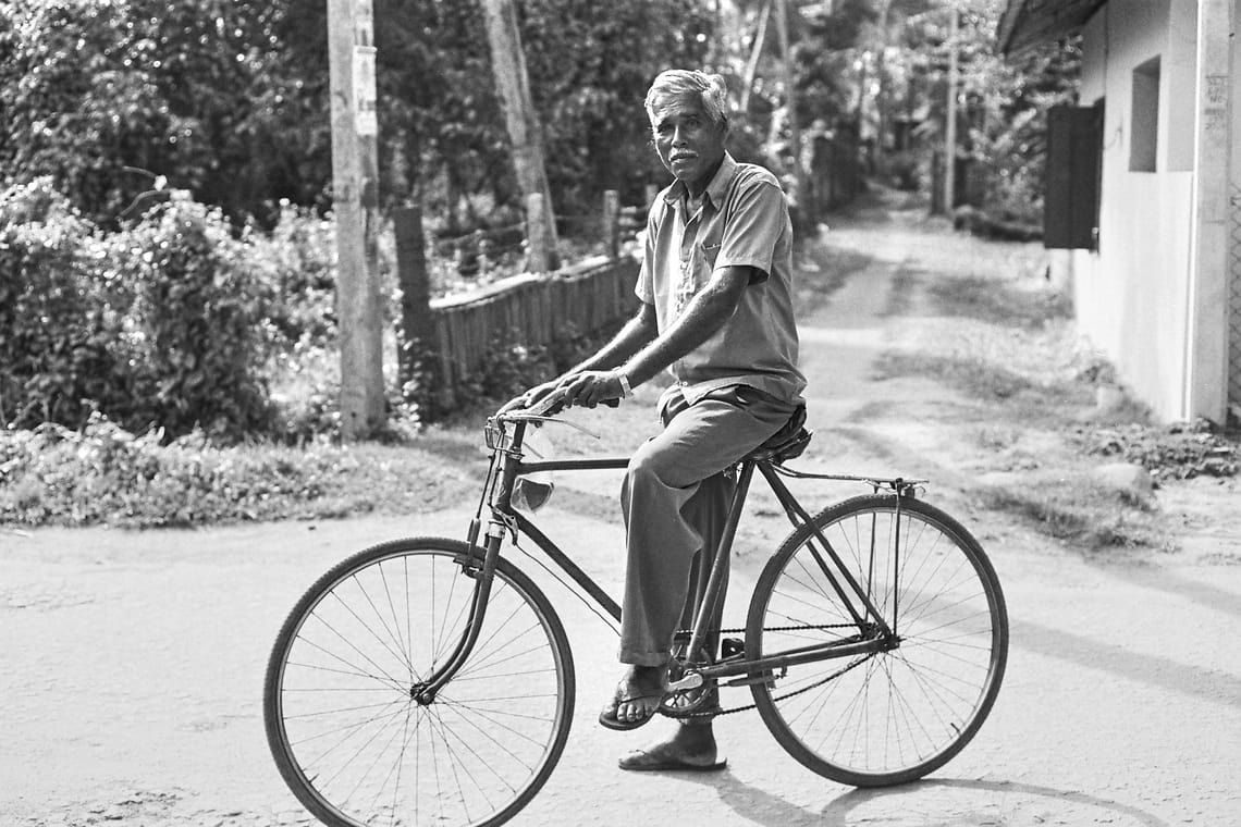 Un homme âgé (Adakkan), cheveux et moustache blancs, pose paisiblement assis sur son vélo dans une rue bordée d'arbres, vêtu simplement, avec des sandales. L'image est en noir et blanc.